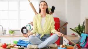 Egy nő meditál a konyhában, miközben a háttérben valaki ételt készít.