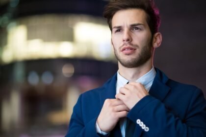 Egy fiatal férfi elegáns öltönyben, nyakkendőt igazítva, komoly arckifejezéssel.