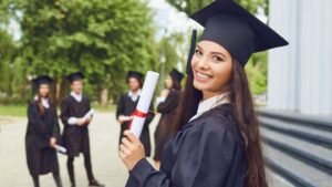 Egy fiatal nő ballagási ruhában mosolyogva tartja a diplomáját, mögötte más ballagók állnak.