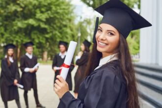 Egy fiatal nő ballagási ruhában mosolyogva tartja a diplomáját, mögötte más ballagók állnak.