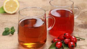 Két csésze csipkebogyótea, az egyik világosabb, a másik sötétebb, friss csipkebogyókkal és citromszelettel a háttérben.