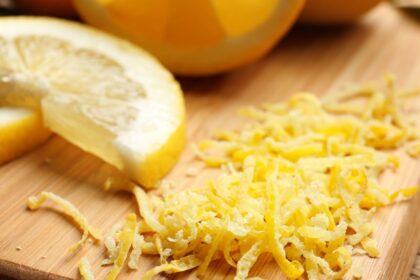 Friss citromhéj reszelve, mellette egy fél citrom, fából készült deszkán.