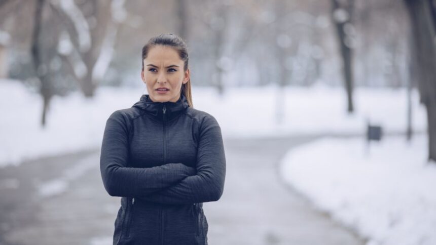 Egy nő sportos öltözetben áll a hóval borított parkban, karba tett kézzel, a hideg időjárás ellenére edzésre készül.