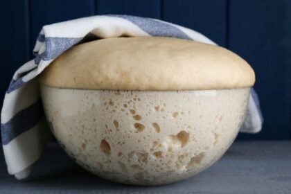 Egy üveg tálban megkelt tészta, amelyet konyharuhával takartak le, a háttérben sötétkék fal.