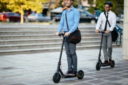 Két férfi elektromos rollerrel közlekedik egy városi téren, vidám hangulatban.