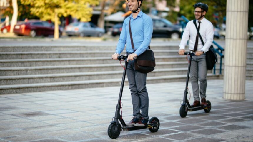 Két férfi elektromos rollerrel közlekedik egy városi téren, vidám hangulatban.