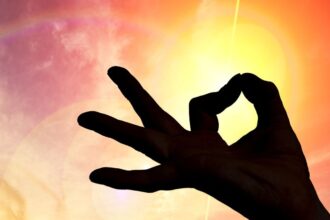 Egy kéz, amely a napfényben meditációs pozíciót mutat, színes égbolttal a háttérben.