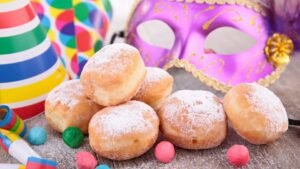 Frissen sült farsangi fánkok porcukorral meghintve, színes farsangi kellékek között.