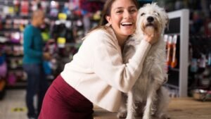 Egy fiatal nő boldogan öleli át a fehér kutyáját egy állatboltban, miközben a háttérben különböző állatfelszerelések láthatók.