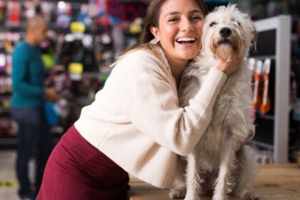 Egy fiatal nő boldogan öleli át a fehér kutyáját egy állatboltban, miközben a háttérben különböző állatfelszerelések láthatók.