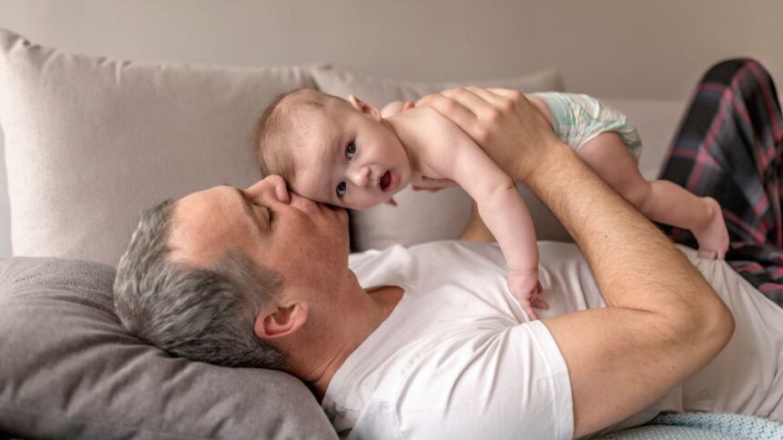 Egy apuka és kisbabája együtt pihennek a kanapén, a férfi szeretetteljesen csókolja a gyermeket.