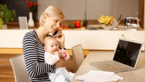 Egy fiatal nő telefonál, miközben a karjában tartja a babáját, a konyhában.