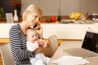 Egy fiatal nő telefonál, miközben a karjában tartja a babáját, a konyhában.