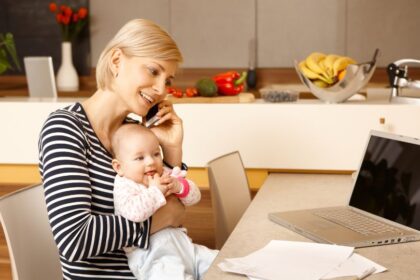 Egy fiatal nő telefonál, miközben a karjában tartja a babáját, a konyhában.