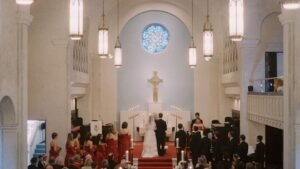 womensway 134 Egy templomi esküvő jelenete, ahol a pár az oltár előtt áll, körülöttük vendégek és koszorúslányok piros ruhákban.