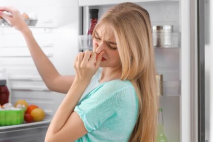 Egy fiatal nő kellemetlen szagot érez a hűtőszekrényből, miközben az ajtaját nyitja.