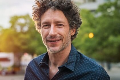 Egy mosolygó férfi áll a városban, zöld fák és lámpák között, barátságos arckifejezéssel.