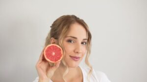 womensway 141 Egy fiatal nő mosolyogva tart a kezében egy fél grapefruitot, ami gazdag vitaminokban és antioxidánsokban, segítve az egészséges bőr fenntartását.