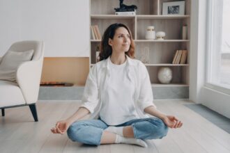 Egy nő meditál a világos, modern lakásban, nyugodt arckifejezéssel.