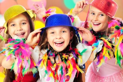 Három vidám gyerek farsangi jelmezben, színes kalapokkal és tollakkal díszítve, boldogan mosolyognak.
