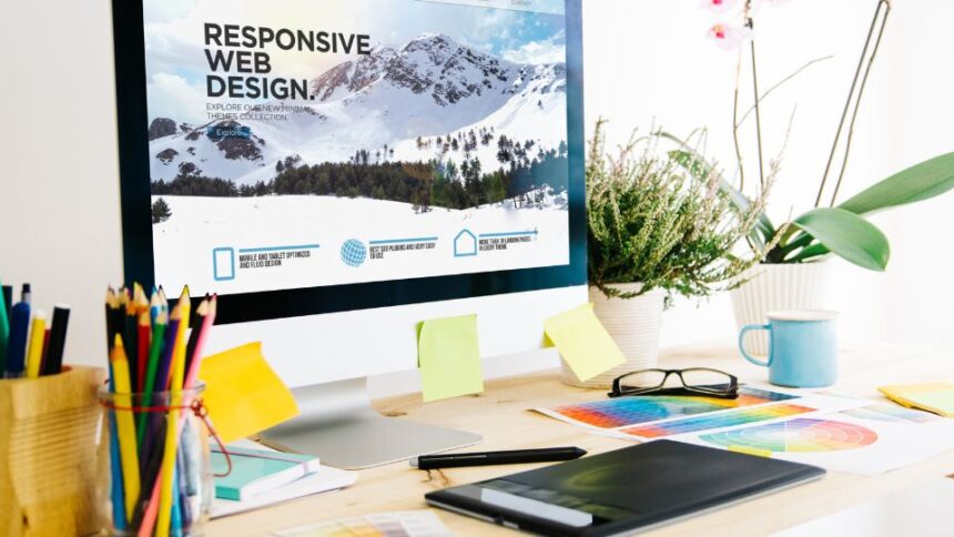 Egy modern munkahely grafikai tervezéshez, ahol egy számítógép képernyőjén responsive webdesign látható, színes ceruzák és tervezési anyagok mellett.