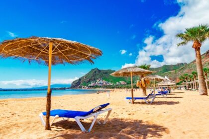 Napernyők és napozóágyak a Kanári-szigetek homokos tengerpartján, háttérben hegyekkel.