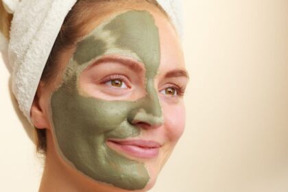 Egy fiatal nő arca zöld arcpakolással, törölközővel a haján, mosolyogva.