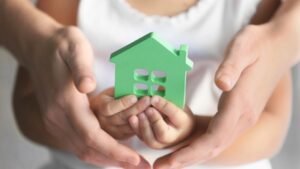 Egy gyermek és felnőttek kezei között egy zöld házikó forma, amely az otthon melegségét szimbolizálja.