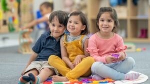 Három kisgyerek ül a szőnyegen, játékokkal körülvéve, boldogan mosolyognak.