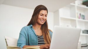 Egy fiatal nő mosolyogva dolgozik a laptopján, otthoni környezetben.