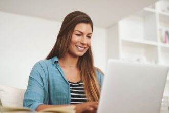 Egy fiatal nő mosolyogva dolgozik a laptopján, otthoni környezetben.