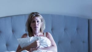 womensway 141 Egy nő rémült arccal ül az ágyban, kezében párnával, sötét szobában.