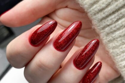 Egy kéz, amelyen csillogó, piros műkörmök láthatók, a háttérben egy világos szövet.