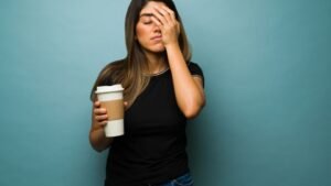 Egy fáradt nő, aki kávét tart a kezében, az arcát a kezébe temeti, jelezve a kimerültséget.