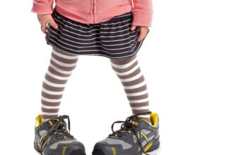 Egy kisgyermek nagy sportcipőben áll, miközben csíkos leggings és szoknya van rajta.