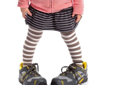 Egy kisgyermek nagy sportcipőben áll, miközben csíkos leggings és szoknya van rajta.