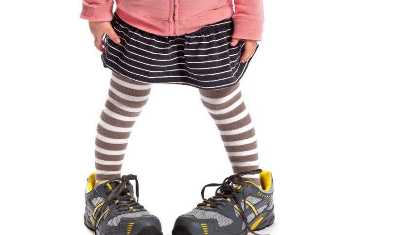Egy kisgyermek nagy sportcipőben áll, miközben csíkos leggings és szoknya van rajta.