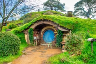Egy varázslatos hobbit ház zöld dombon, kék ajtóval és virágokkal.