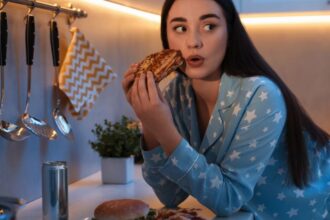 Egy fiatal nő pizsamában, egy falat pizzát tart a kezében, miközben a konyhában ül.