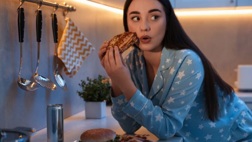 Egy fiatal nő pizsamában, egy falat pizzát tart a kezében, miközben a konyhában ül.