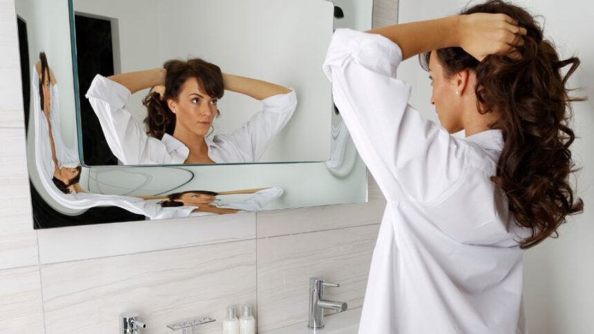 10 tipp, hogy gyorsan elkészülj reggelente 1 Egy fiatal nő a tükör előtt áll, miközben a haját fésüli reggel. Fehér ingben, koncentrált arckifejezéssel készülődik a napra.