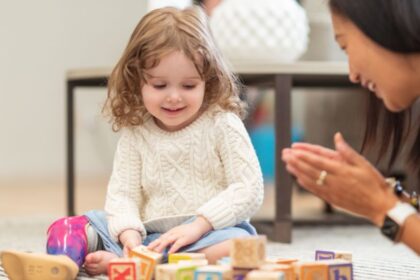 Egy kisgyermek játszik színes építőkockákkal, miközben egy felnőtt figyeli és támogatja őt.