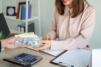 Egy nő mosolyogva fogad pénzt egy férfi kezéből az irodában, ahol számológép és iratok találhatók.
