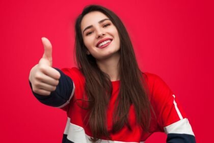 Egy fiatal nő mosolyogva mutatja a hüvelykujját felfelé, piros háttér előtt.