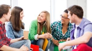 Öt fiatal barát beszélget egy világos szobában, mosolyogva és nevetve, miközben könyveket és jegyzeteket tartanak a kezükben.