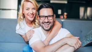 womensway 142 Egy boldog pár ölelkezik a kanapén, mosolyogva néznek a kamerába, ami a szeretetteljes kapcsolatot tükrözi.