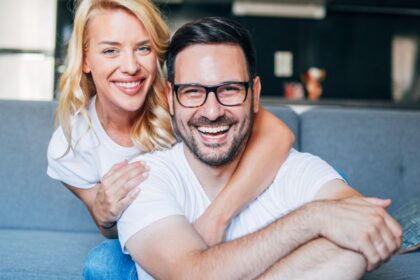 Egy boldog pár ölelkezik a kanapén, mosolyogva néznek a kamerába, ami a szeretetteljes kapcsolatot tükrözi.