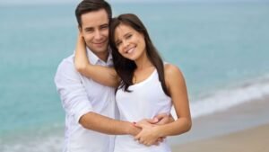 Egy fiatal pár ölelkezik a tengerparton, boldogan mosolyognak egymásra.