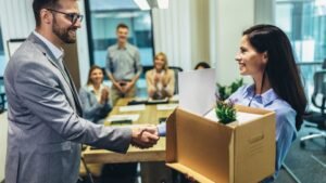 womensway 135 Egy fiatal nő mosolyogva kézfogással üdvözli új főnökét, miközben egy dobozt tart a kezében, mögöttük kollégák tapsolnak.