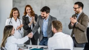 Egy új munkatárs üdvözlése a munkahelyen, ahol a csapat tagjai örömmel tapsolnak és gratulálnak.